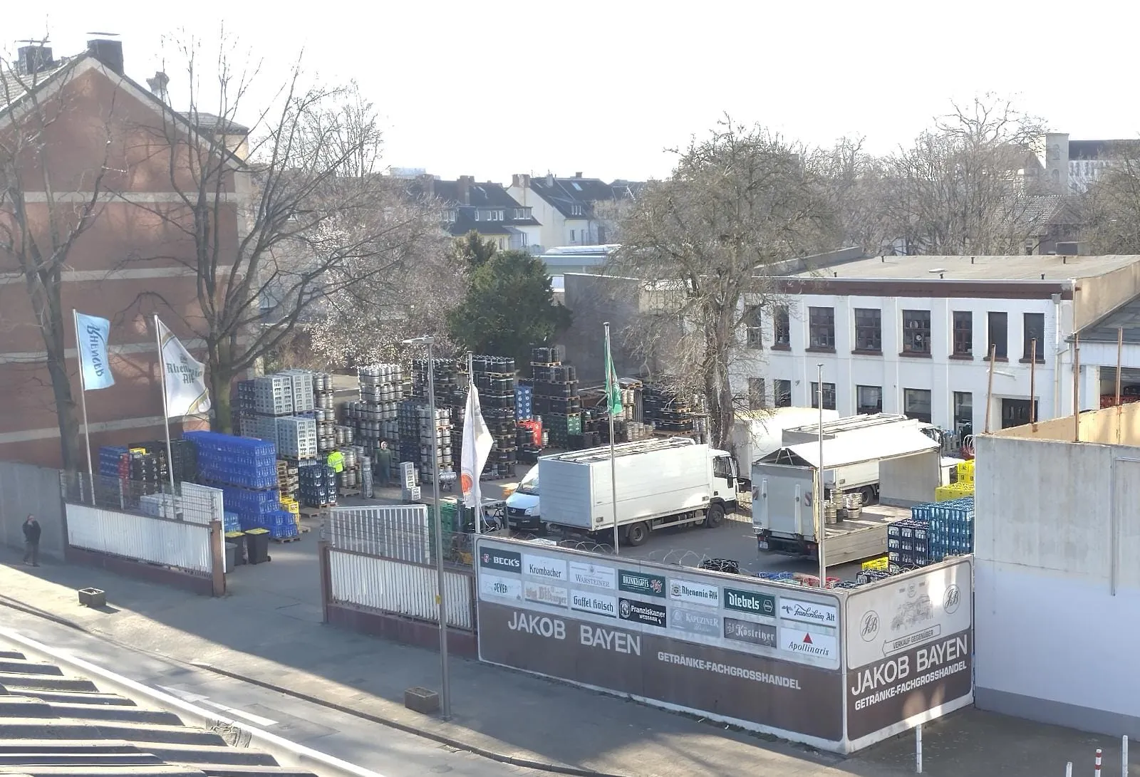 The yard of Jakob Bayen KG in Krefeld seen from above: stacked beverage crates and kegs, delivery trucks, and the company sign "Jakob Bayen Getränke-Fachgrosshandel".